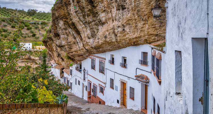 White houses tucked beneath an immense overhanging cliff face along a cobbled lane.