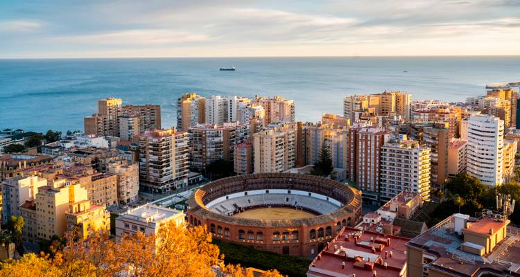 Cityscape of Malaga with round bullring arena, autumn trees and Mediterranean Sea at sunset.