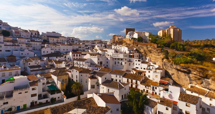 Whitewashed houses of Setenil de las Bodegas cascade down rocky hillside under bright blue sky.