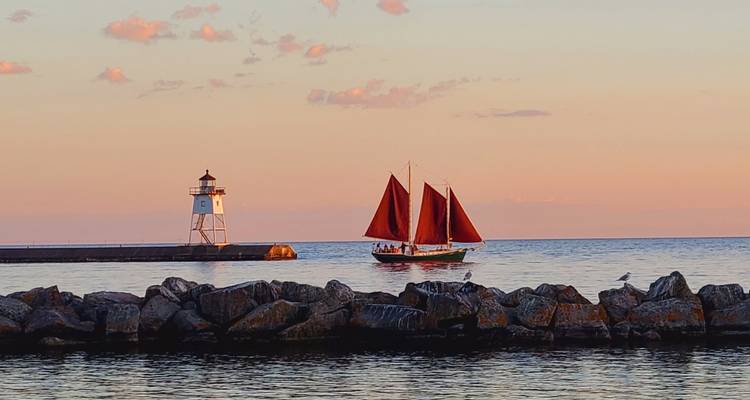 Goélette aux voiles rouges passant devant un phare au coucher de soleil aux teintes pastel sur le lac Supérieur