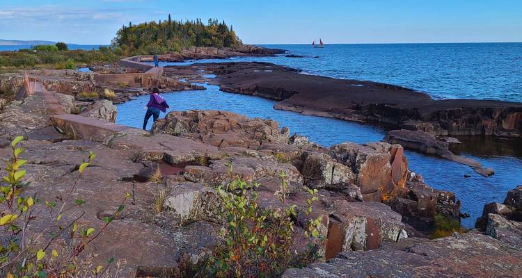 Randonneur sur un littoral volcanique accidenté regardant vers un voilier distant et un lac bleu