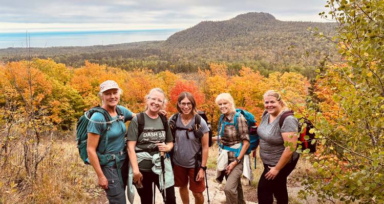 Cinq randonneurs souriants posant sur un sentier avec un feuillage automnal vibrant et une crête montagneuse en arrière-plan