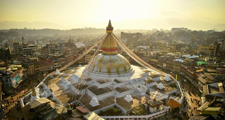 Gouden-uur luchtfoto van de enorme witte Boudhanath Stupa gekroond met wapperende gebedsvlaggen, omgeven door de stad en mistige voethellingen.