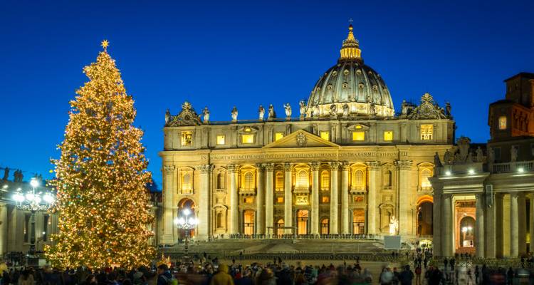 Fachada iluminada de la Basílica de San Pedro y un enorme árbol de Navidad brillando contra un profundo cielo nocturno azul.