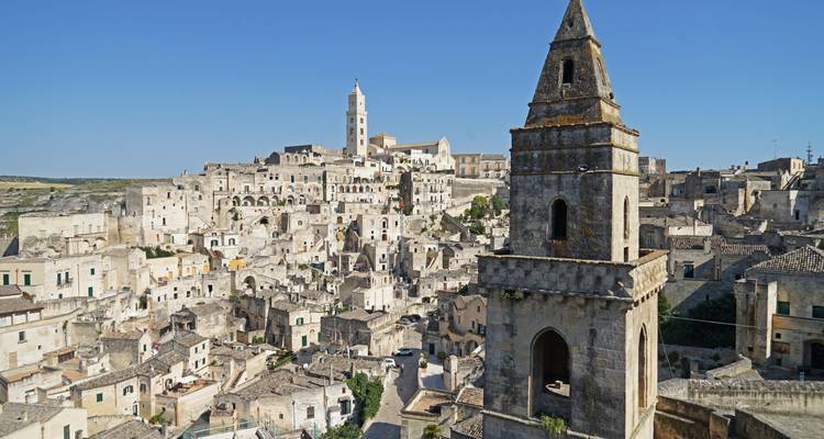 Panorámica diurna de las viviendas de piedra caliza de Matera y su alta torre de iglesia bajo un cielo azul despejado.