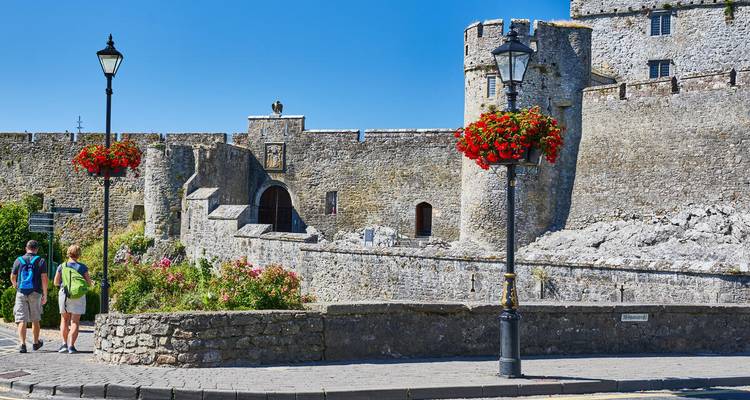Los mochileros se acercan a los muros de piedra gris y torres del Castillo de Cahir adornados con cestas de flores colgantes