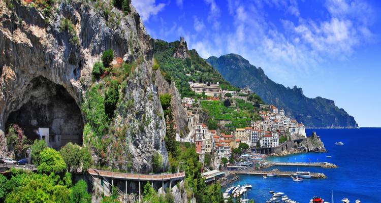 Panoramisch uitzicht op het stadje Amalfi dat tegen groene rotsen aan hangt boven een glitterend kobaltblauw zee en jachthaven.