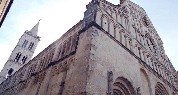 Angle ascendant de la facade en pierre de la Cathédrale de Zadar et de son campanile affichant des arcs romans et une rosace contre un ciel lumineux