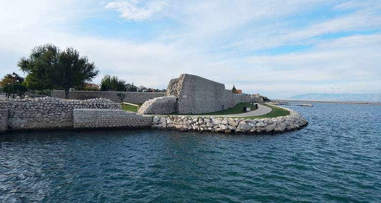 Mur de fortification en pierre s'incurvant le long d'une baie turquoise avec des pelouses entretenues et des montagnes lointaines.