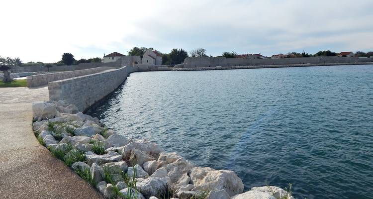 Long mur de mer bordant des eaux côtières calmes avec des enrochements et des maisons villageoises au loin.