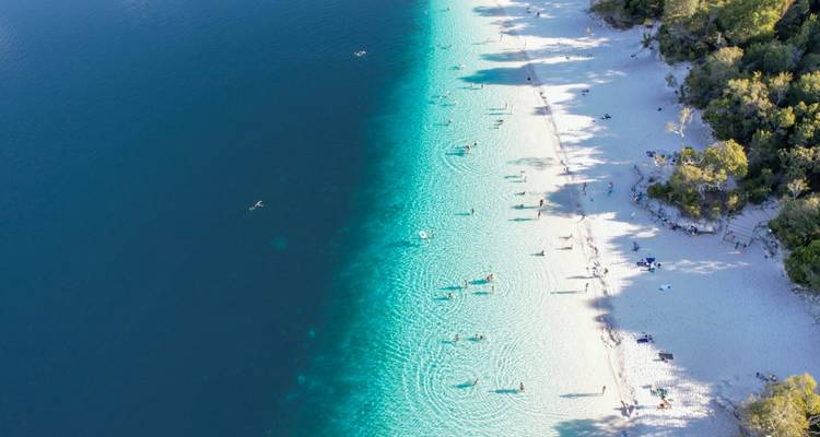 Stunning aerial of a crystal-clear lake with bright white sandy shore dotted with swimmers.