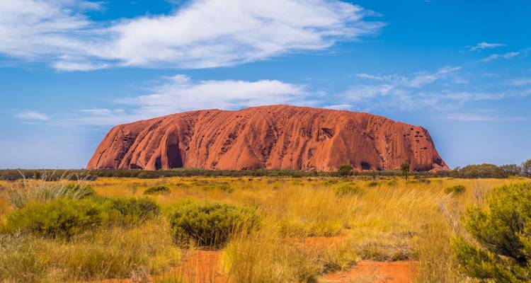 Iconic red sandstone monolith Uluru rising from flat desert grassland under a blue sky.