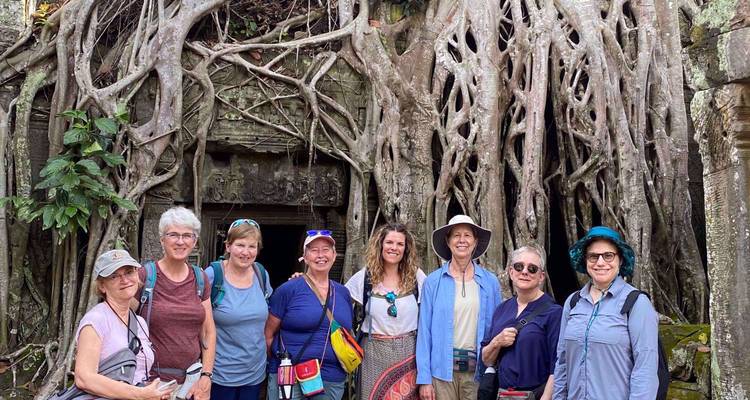 Lächelnde Reisegruppe posiert vor massiven Baumwurzeln, die den Steintempelingang von Ta Prohm umhüllen