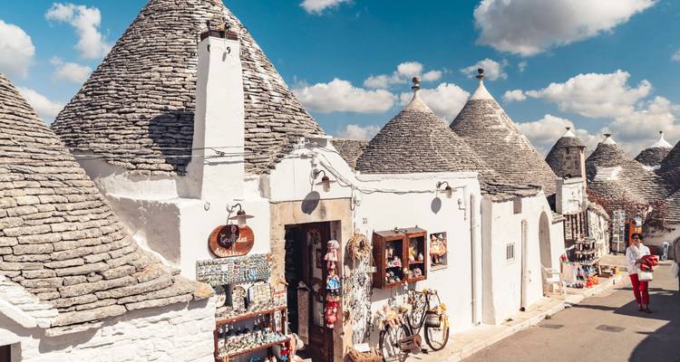 Les maisons trulli blanchies à la chaux traditionnelles aux toits en pierre conique bordent une rue ensoleillée sous un ciel bleu avec quelques nuages, mettant en avant les artisanats locaux exposés à l'extérieur.