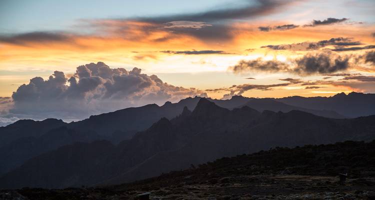 Les silhouettes colorées du coucher de soleil épousent les crêtes montagneuses dentelées avec des nuages dramatiques au-dessus.