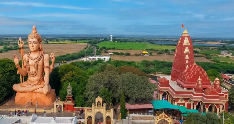 Gigantische sitzende Shiva-Statue neben einem Tempel aus rotem Backstein inmitten grüner Felder