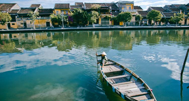 Traditional wooden boat with conical-hat rower reflects in calm canal of Hoi An ancient town