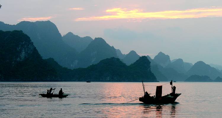 Silhouetten von Fischerbooten treiben bei Sonnenaufgang in Pastelltönen über ruhige Gewässer in der Halong-Bucht.