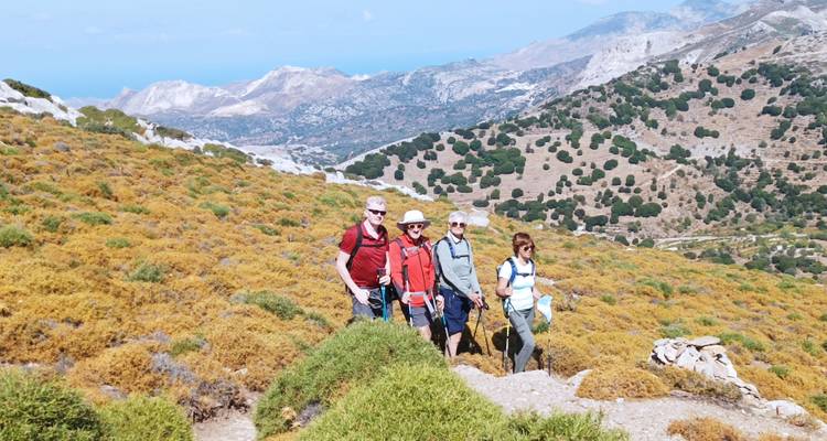Cuatro excursionistas posando en un sendero de ladera bañado por el sol con vistas panorámicas de islas griegas y pendientes escarpadas detrás de ellos.
