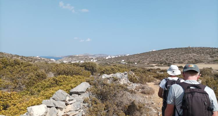 Dos mochileros caminan por un sendero rocoso en una isla rodeado de arbustos bajos con pueblos blancos distantes visibles hacia el mar.