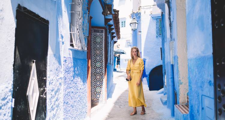 Vrouw in gele jurk staande in een zonnige blauwe straat van Chefchaouen