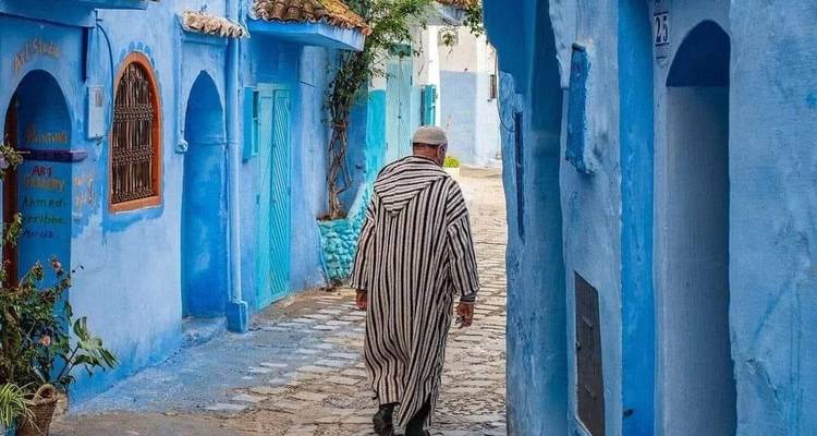 Man in a gestreepte djellaba die door een smal blauw geverfd steegje in Chefchaouen loopt.