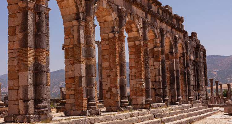 Zuilengalerij van verweatherde stenen bogen uit de Romeinse ruïnes van Volubilis met bergen op de achtergrond.