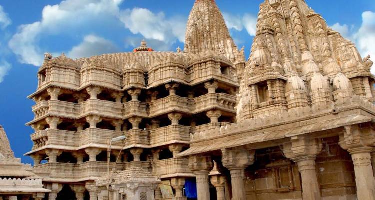 Eine kunstvoll geschnitzte Fassade eines Jain-Tempels zeigt mehrstöckige Balkone unter einem hellen Himmel mit Wolkenformationen.