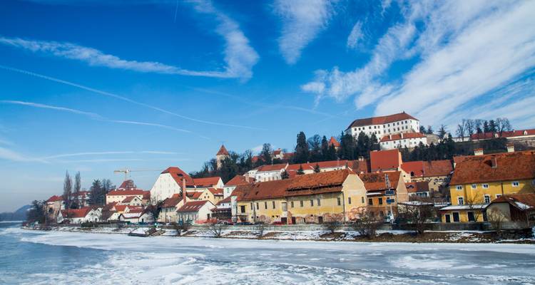 Ville riveraine couverte de neige avec des toits rouges et un château au sommet de la colline sous un ciel bleu strié.