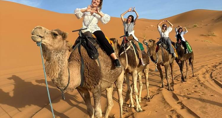 Vrouwen rijden op kamelen over gigantische oranje zandduinen en maken hartjes met hun handen onder een blauwe lucht.