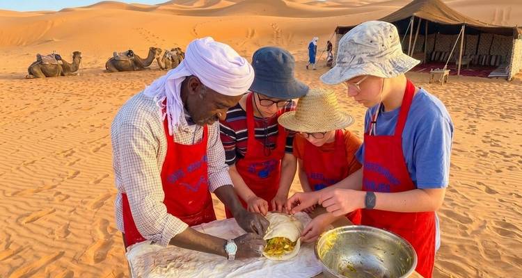 Reizigers met schorten leren traditioneel eten klaar te maken op woestijnzand met kamelen op de achtergrond.