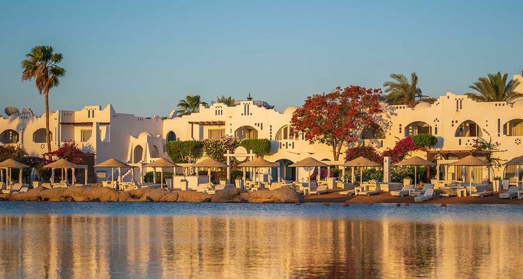 Resort en bord de plage aux murs blanchis à la chaux avec des arbres en fleurs se reflétant dans une eau calme sous une lumière de lever de soleil chaleureux.