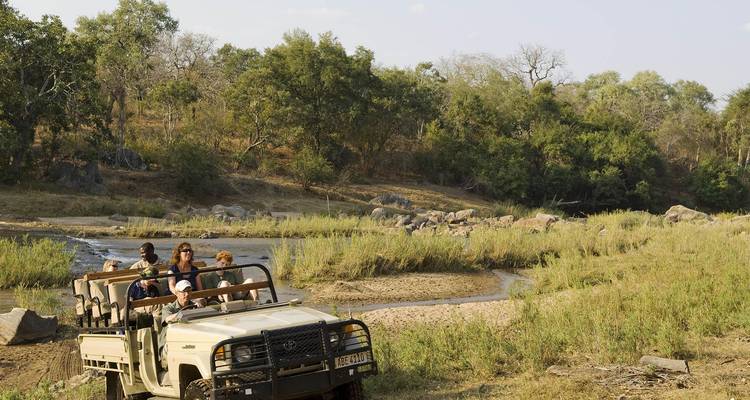 Open safari vehicle with tourists drives beside a winding river in bushland dotted with trees and rocks.