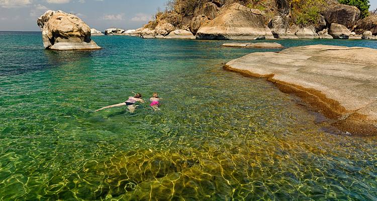 Two snorkellers swim in crystal-clear emerald water beside smooth granite boulders on a lake shore.