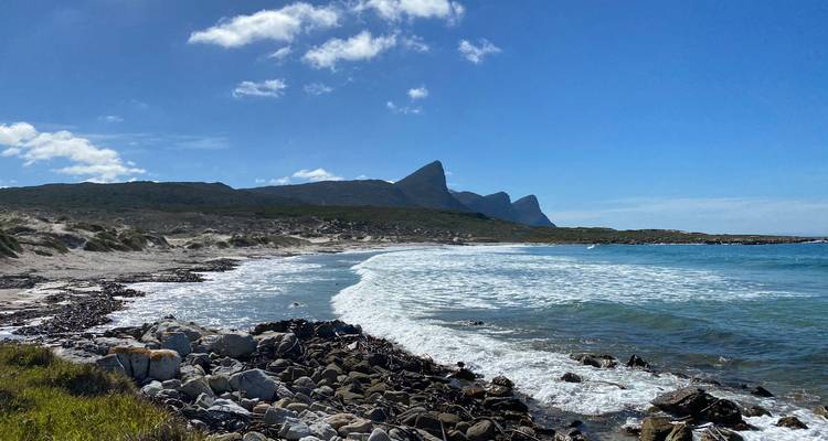Rocky beach with rolling surf and dramatic jagged peaks rising in the distance under a clear blue sky.