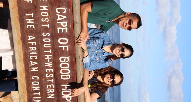 Three travellers smile behind the wooden Cape of Good Hope sign with ocean waves in the background.