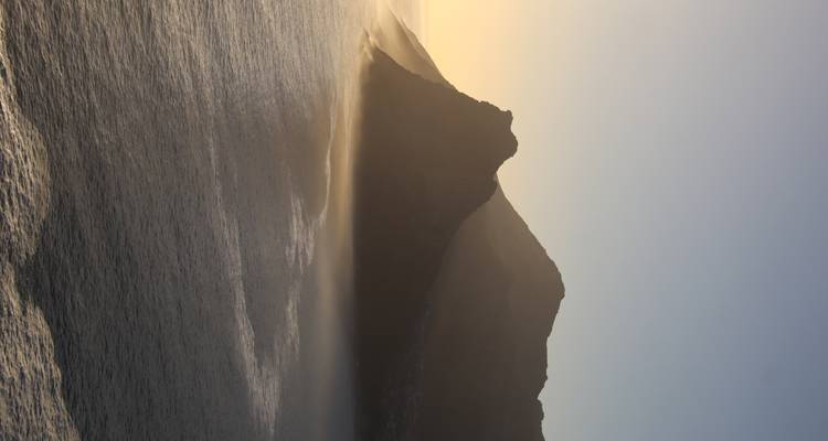 Golden light bathes rugged coastal mountains with mist rising from the ocean at dusk.