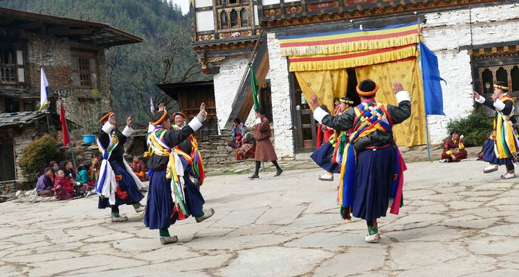 Bailarines butaneses con fajas y sombreros coloridos realizan un baile en círculo en el patio de un monasterio mientras los aldeanos los observan.