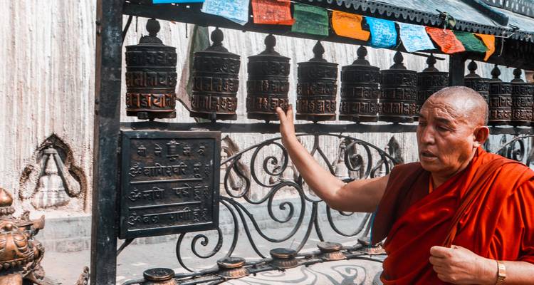 Monje con túnica roja girando una fila de ruedas de oración ornamentadas bajo banderas de oración que ondean.