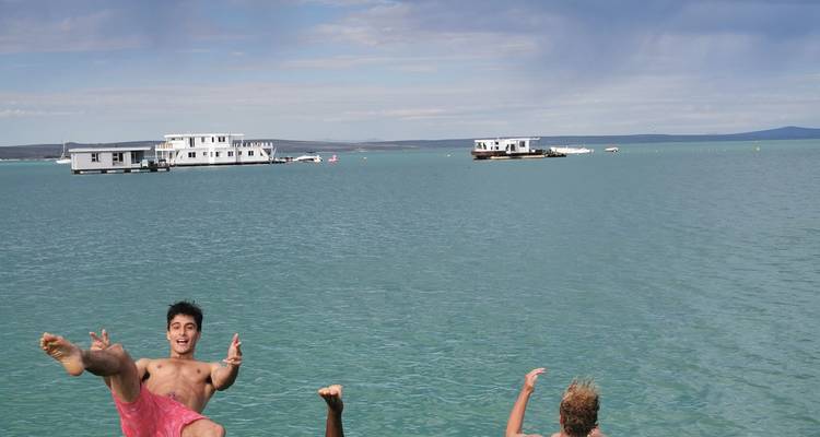 Young travellers jump gleefully into a calm turquoise lagoon with houseboats anchored in the background.