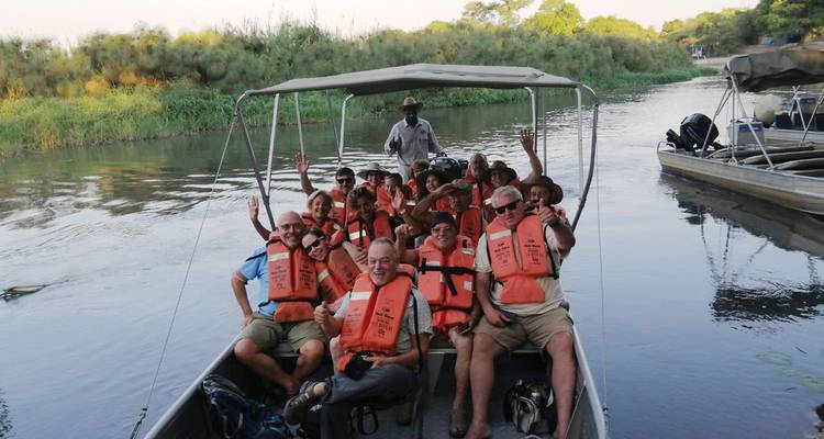 Un groupe de touristes souriants portant des gilets de sauvetage fait signe depuis un petit bateau de safari sur le fleuve Chobe.