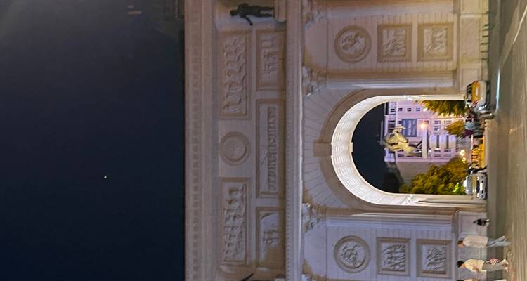 Vista nocturna del arco del triunfo iluminado con peatones y autos estacionados.