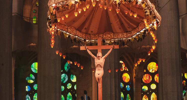 Crucifijo central y dosel iluminado dentro de la Sagrada Família rodeado de ventanas coloridas.