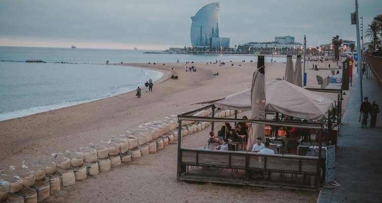 Vista nublada a última hora del día de la Playa de Barceloneta con una terraza de cafetería frente al mar y el hotel W Barcelona con forma de vela que domina el tranquilo Mediterráneo.
