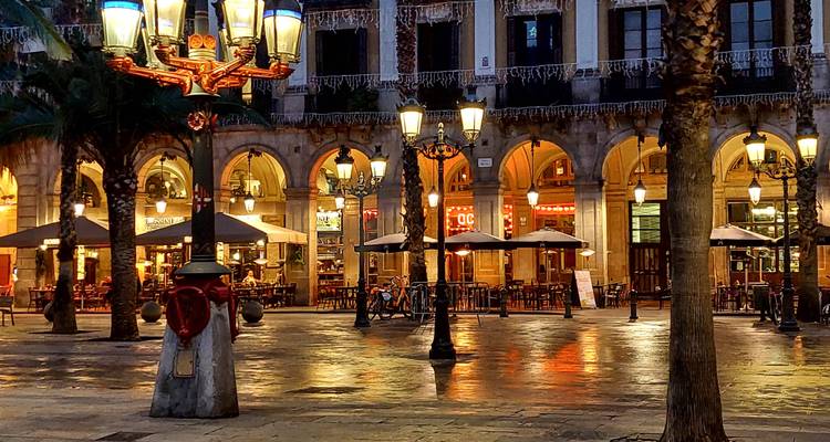 Escena nocturna de una plaza con arcadas rodeada de farolas brillantes y cafés al aire libre, con piedra mojada reflejando las luces cálidas.