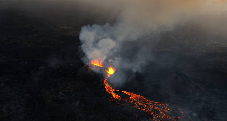 Luftaufnahme eines ausbrechenden Vulkans mit glühenden Lavafluss und aufwallender Rauchentwicklung.