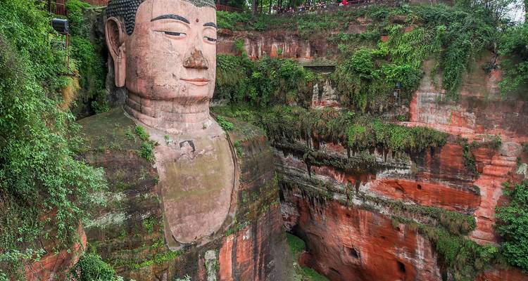 Le massif Bouddha géant de Leshan, sculpté dans une paroi rocheuse entouré d'une végétation luxuriante et de murs de grès rouge.