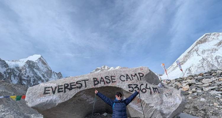 El trekero levanta los bastones triunfalmente junto a una roca en el Campamento Base del Everest con picos nevados de fondo