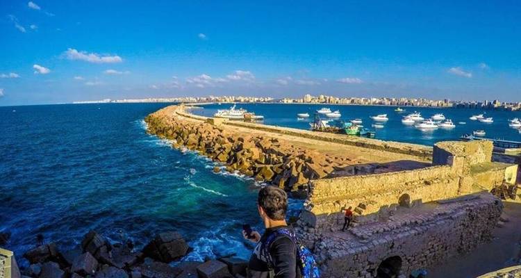 Un viajero con una mochila mira hacia un muelle de piedra lleno de botes en el vibrante mar Mediterráneo azul.