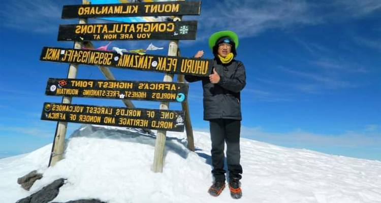Een triomfantelijke klimmer poseert naast het Uhuru Peak-bord bovenop de sneeuwige Mount Kilimanjaro onder een diepblauwe hemel.